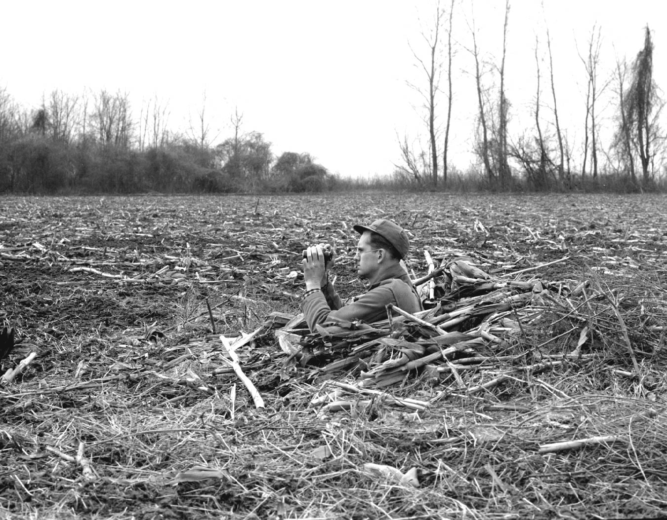 Man in field, in hole up to his chest, holding binoculars. 